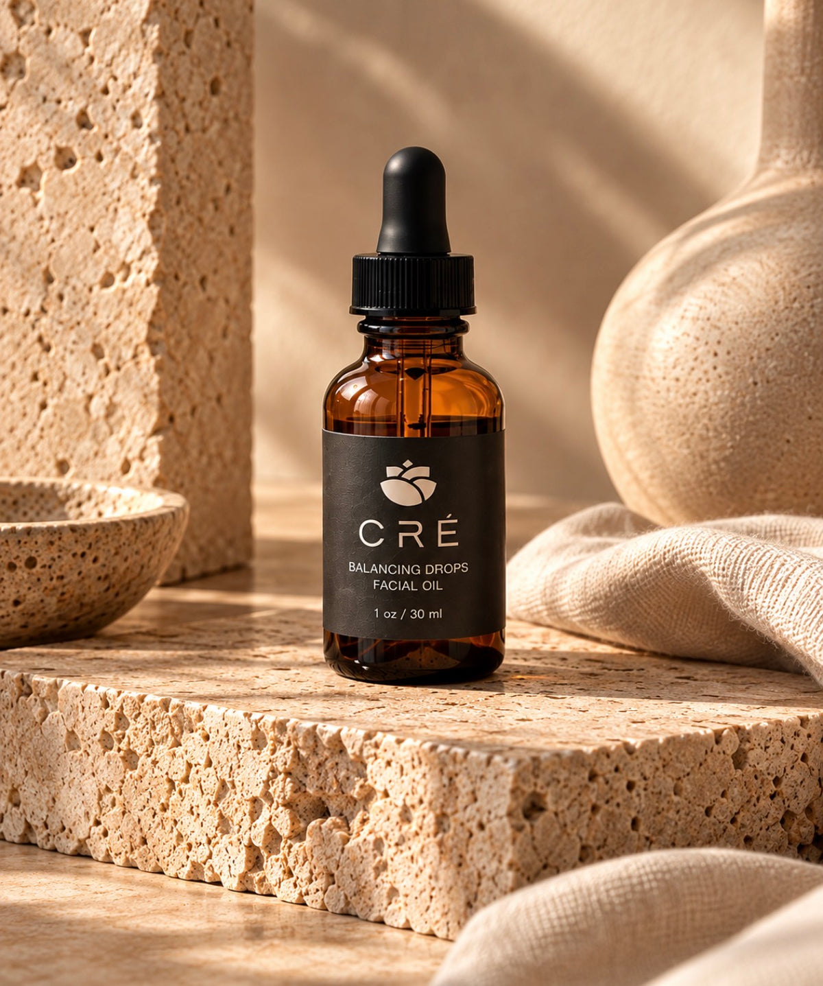 An amber glass dropper bottle of CRÉ Balancing Drops Facial Oil stands on a textured stone slab with neutral beige props in a warm, sunlit setting.