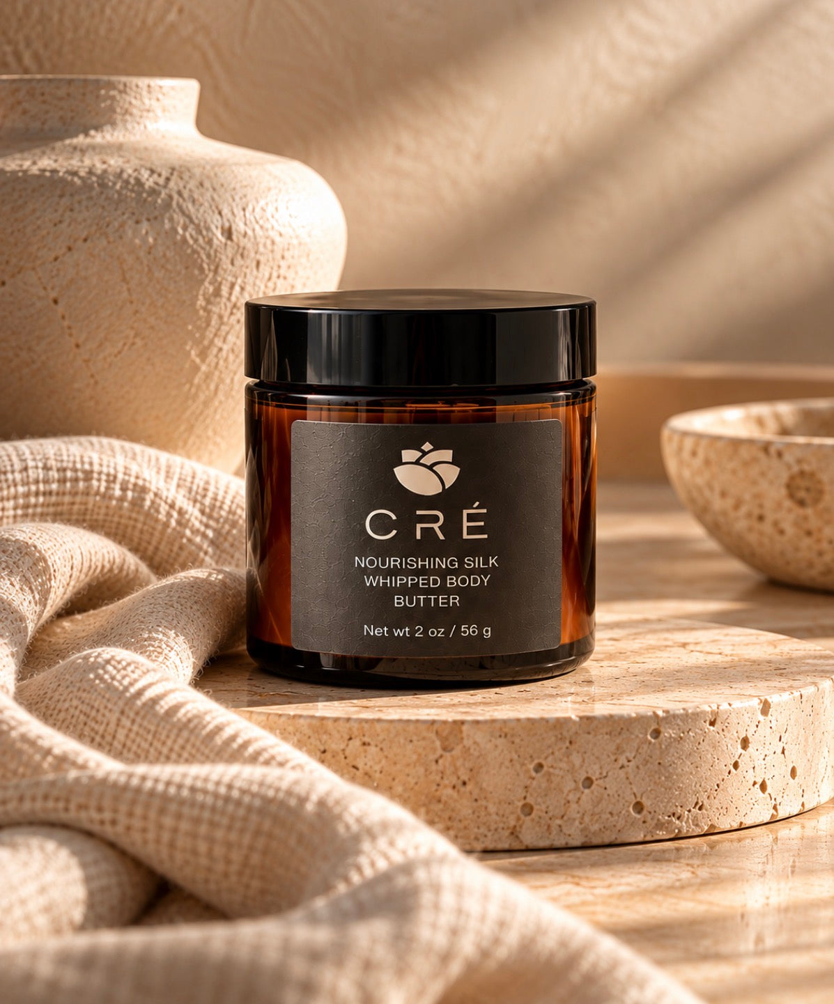Crè Nourishing Silk Whipped Body Butter in an amber jar with black lid, styled on warm neutral stone and linen.